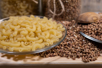 Buckwheat is scattered on a wooden board. Nearby is a transparent plate with pasta.