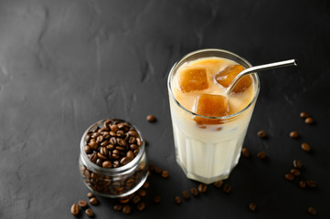 Transparent glass with milk and ice cubes made from coffee. Iced coffee with a metal tube and grains in a jar on a dark background. Top view with copy space.