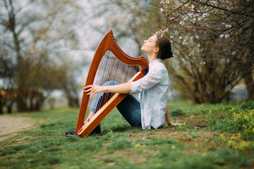 Woman harpist sits on grass and plays harp among blooming apricot trees.