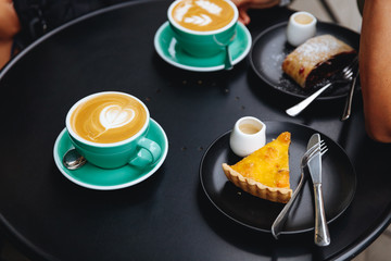 Hot and tasty cappuccino in two turquoise colored cups with delicious strudel and cheesecake placed on black round table of coffee shop terrace. Sweet break during everyday routine.
