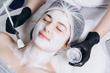 Young woman having spa procedure on her face. Woman lying on spa table and cosmetologist applying cosmetic mask on the her face in sap salon. Copy space for your text