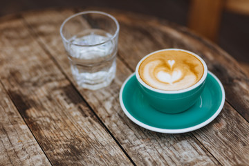 Beautiful latte art in turquoise colored ceramic cup with glass of water on wooden table with vintage effect. Hot coffee with lush milk foam served for one person outdoors.