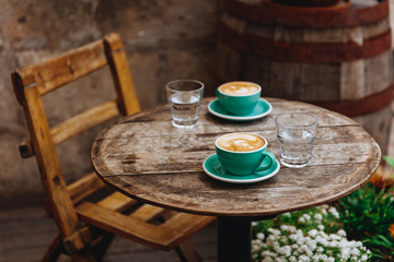Beautiful cafe terrace with wooden chair and table and greenery ceramic cups of tasty aroma cappuccino with heart shaped milk foam. Morning hot beverage served with glass of water.
