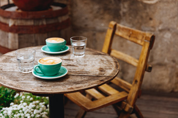 Morning aroma cappuccino with lush milk foam on top in two turquoise colored ceramic cups with glasses of water on round wooden table of coffee shop terrace. Comfortable place for breakfast