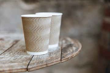 Close up of two disposable brown cups of hot flavored coffee on rustic wooden table of coffee shop terrace. Vintage effect and blur background