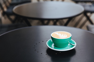 Black round table with trendy colored cup of freshly brewed aroma coffee with latte art outside. Morning hot beverage served on beautiful terrace of coffee shop