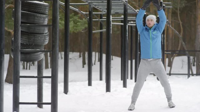 Medium shot with slowmo of active man in blue windbreaker warming up with jumping jacks and jogging in place while working out at outdoor gym in winter
