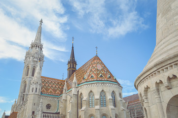 Fototapeta na wymiar The spire of the famous Matthias Church in Budapest, Hungary. Roman Catholic church built in the Gothic style. Orange colored tile roof. Blue sky and white clouds above. Horizontal photo with filter