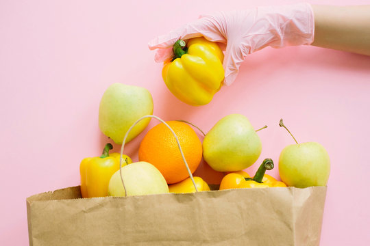 Hand In Pink Glove Putting Yellow Pepper In Paper Bag With Groceries On Pink Background Flat Lay. Safe Shopping In Quarantine. Order Food Online With Delivery And Stay Home. Prevention Of Virus