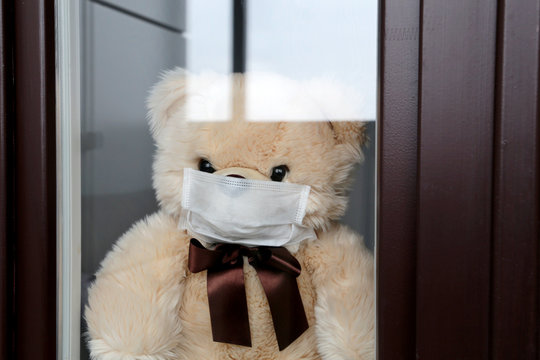Toy bear in a face medical mask looks out of the window. Concept of coronavirus quarantine during the COVID-19 epidemic