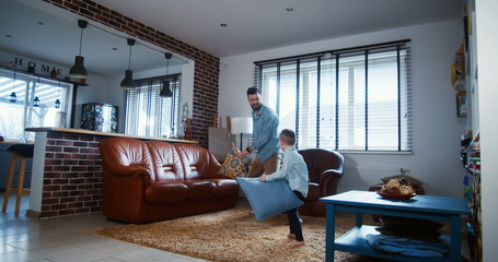 Happy young Caucasian father and little son playing, fighting pillows in modern light house living room slow motion.