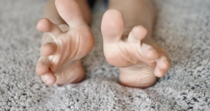 Soles of two feet while someone lays on a fuzzy light grey blanket stretching their toes then a cat comes over and sniffs