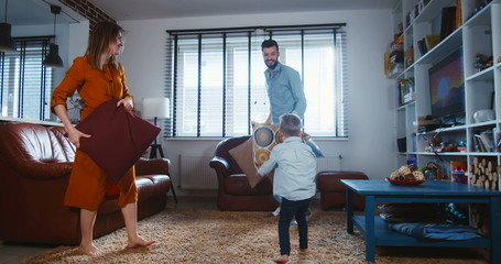 Family fun concept. Happy young European father and mother fight pillows with cute little son in modern home slow motion
