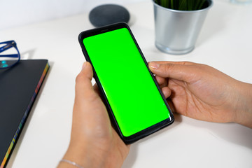 Close up of a Caucasian woman hands holding her mockup phone vertically in a desk with glasses, note, plant and smart assistant