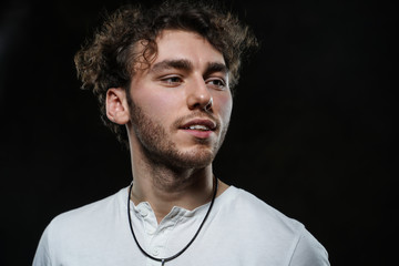Curly and hansome caucasian man standing in a dark studio on a grey background, wearing casual white shirt looking interested