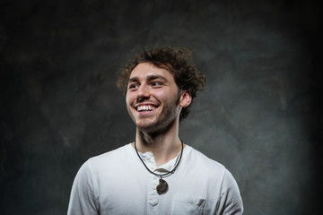 Curly and hansome caucasian man standing in a dark studio on a grey background, wearing casual white shirt looking joyful