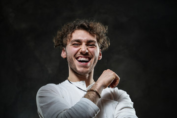 Curly and hansome caucasian man standing in a dark studio on a grey background, wearing casual white shirt looking joyful and positive