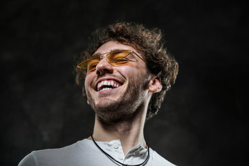 Smiling and cheerful young adult male model posing for a photoshoot on a grey background in a dark studio, wearing white casual shirt and sunglasses while laughing
