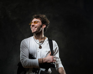 Curly and smiling caucasian male musician standing in a dark studio on a grey background, wearing casual white shirt and glasses while playing an electric guitar.