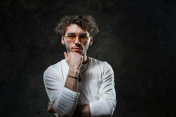 Grumpy looking young adult caucasian man standing in a thinking pose in a dark studio on a grey background, wearing a white casual shirt and sunglasses