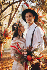 stylish couple of brides: man in tuxedo and hat, woman in light white dress with wreath on head against evening park background, lovers posing at wedding photo shoot. wedding background