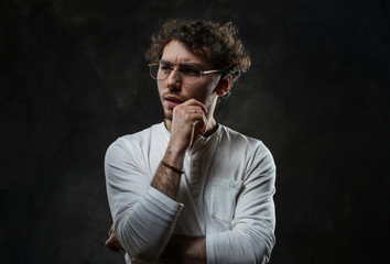 Thoughtful looking young adult caucasian man posing in a dark studio on a grey background, wearing a white casual shirt and glasses