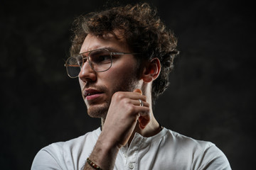 Grumpy looking young adult caucasian man posing in a dark studio on a grey background, wearing a white casual shirt and glasses
