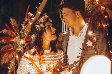 Stylish couple of brides, pretty woman in wedding dress and young man in fedora hat. Romantic ceremony in nature. The lights of the electric garland illuminate the wedding party. Wedding background