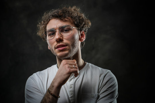 Sceptically looking young adult caucasian man posing in a dark studio on a grey background, wearing a white casual shirt and glasses