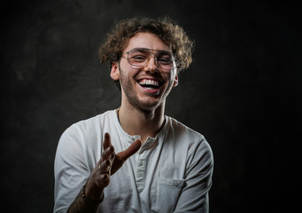 Smiling and cheerful young adult male model posing for a photoshoot on a grey background in a dark studio, wearing white casual shirt and glasses, while making a gesture with his hand