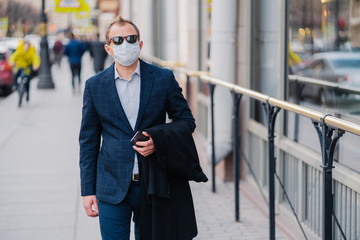 Outdoor shot of businessman in formal clothes, holds modern cell phone in hands, walks on street, protects himself with medical mask, prevents transmissible infectious disease or air pollution in city