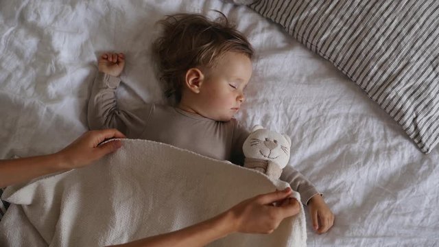 Little baby girl sweetly sleeps in bed, Mother covering with blanket her adorable sleeping daughter in bedroom