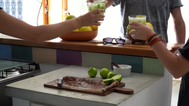 Three friends hands celebrating and toasting glasses with Brazilian Caipirinha drink, made with cacha&ccedil;a and lemon at home during sunny summer day. Friendship, happiness, celebration concept.