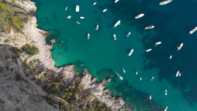Aerial drone top view tracking shot of huge number of luxury boats berth over crystal clear seawater in Capri Italy near rock cliffs with colorful trees
