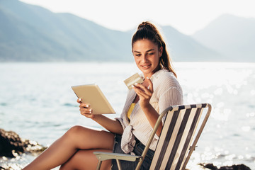 Fototapeta premium Smiling woman sitting on deck chair by the sea using tablet and credit card on a sunny day