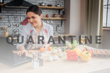 Cheerful adult woman sit at table in kitchen. She typing on keyboard and look on screen. Woman cook...