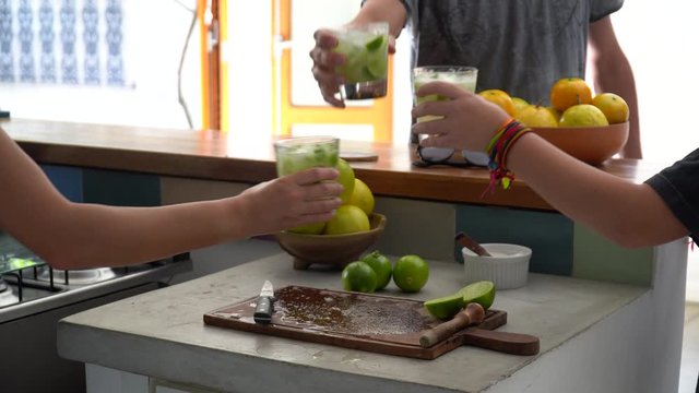 Three friends hands celebrating and toasting glasses with Brazilian Caipirinha drink, made with cacha&ccedil;a and lemon at home during sunny summer day. Friendship, happiness, celebration concept.