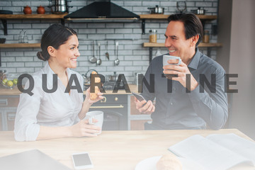 Happy couple sit at table in kitchen. They look at each other. Woman hold croissan and cup in hand. Guy has phone. Happy people smile.