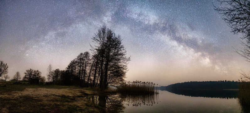 beautiful natural panoramic photo of the milky way over a lake and a tree on a beach, reflections in the water and two shooting stars flying in the night sky  
