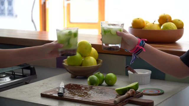 Three friends hands celebrating and toasting glasses with Brazilian Caipirinha drink, made with cacha&ccedil;a and lemon at home during sunny summer day. Friendship, happiness, celebration concept.