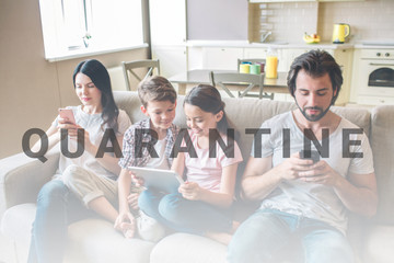 Parents are sitting on sofa with kids and look at phones. Children are in bettween woman and man. Girl holds tablet in hands. They look at screen.