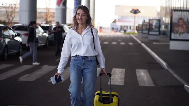 Excited, stylish woman walk with trolley yellow case by empty airport terminal outside road, slender female wearing jeans and white shirt. Happy, smilling woman walking to take her plane with passport