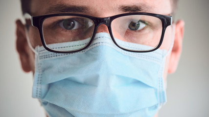 Close-up portrait of young sad and tired doctor in misted glasses and medical mask looking into camera