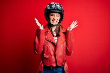 Young beautiful brunette motocyclist woman wearing motorcycle helmet and red jacket celebrating mad and crazy for success with arms raised and closed eyes screaming excited. Winner concept