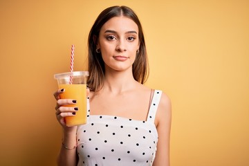 Young beautiful brunette woman drinking healthy orange juice over yellow background with a confident expression on smart face thinking serious