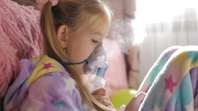 Little girl holds inhaler mask at home. Sick kid breathes through a nebulizer. Baby using equipment to treat asthma or bronchitis. Home treatment.