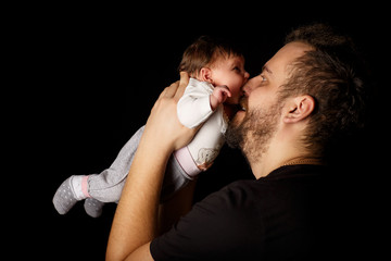 a bearded father in a black T-shirt holds a newborn baby. the child bites his father’s nose