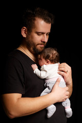 a bearded father in a black t-shirt holds a newborn baby. Dad kisses and hugs daughter or son on a black background