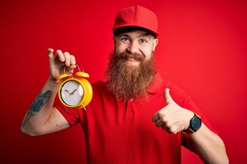 Redhead Irish delivery man with beard holding alarm clock for deadline over red background happy with big smile doing ok sign, thumb up with fingers, excellent sign