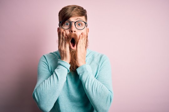 Handsome Irish redhead man with beard wearing glasses over pink isolated background afraid and shocked, surprise and amazed expression with hands on face